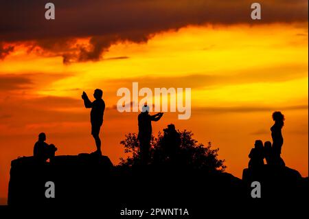 Silhouetten von Menschen auf dem Felsen, die Fotos und Selfies vor dem Hintergrund des orangefarbenen Himmels machen Stockfoto