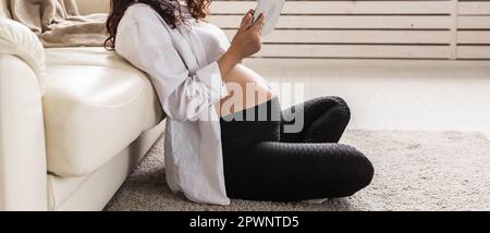 Eine Schwangerin hält das Tablet auf einem Teppich in der Nähe einer Couch im Wohnzimmer zu Hause. Stockfoto