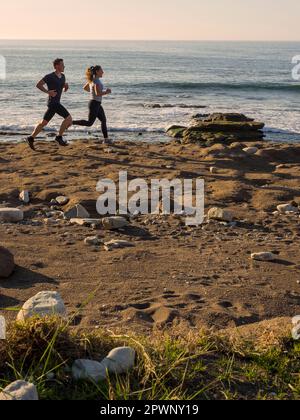 Mann und Frau laufen auf einem einzigen Pfad an der Küste von Azkorri Beach Stockfoto