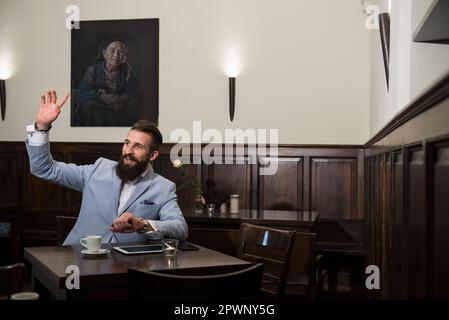 Junger Mann im Anzug ruft Kellner im Restaurant, während Tablet und Kaffeetasse mit Handy auf dem Esstisch standen Stockfoto