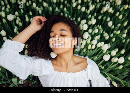 Nahaufnahme des weiblichen Porträts eines süßen, charmanten afrikanischen Mädchens mit sanft weißen Tulpenknospen. Romantisches, junges Afro-Mädchen in einem weißen Hemd Stockfoto