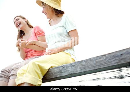 Sie lachten über den See. Eine Mutter und eine Tochter lachten zusammen, während sie auf einem Steg am See saßen. Stockfoto