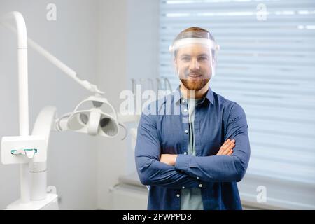 Medizinische Sicherheit. Porträt eines jungen Zahnarztes in der Gesichtsschildposition am Arbeitsplatz in der modernen Klinik. Der Zahnarzt sieht sich die Kamera an, die ihm die Arme kreuzte Stockfoto