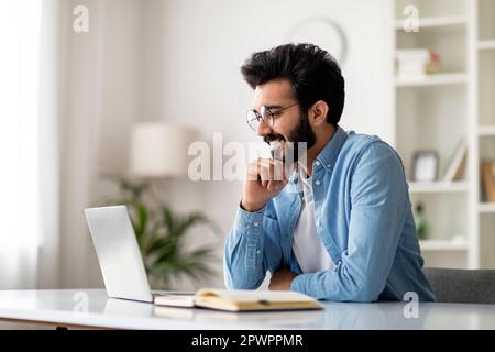 Ein Hübscher Indischer Schriftsteller Sitzt Am Schreibtisch Mit Laptop Und Lächelt Stockfoto