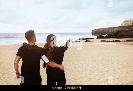 Sportlicher Mann und sportliche Frau, die sich umarmen, eine Trainingspause genießen, die Dame zeigt mit dem Finger auf leere Flächen am Meeresstrand, draußen Stockfoto