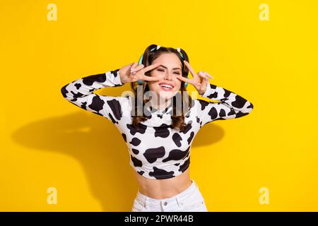 Foto eines positiven Mädchens im gepunkteten Outfit Make V Sign Dancing Hören Headset Melodie isoliert glänzende Farbe Hintergrund Stockfoto