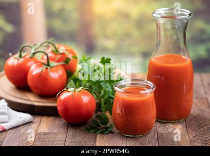 Tomatensaft in Glas und Karaffe mit frischen Tomaten und Petersilie auf Holztisch mit Sommerhintergrund Stockfoto