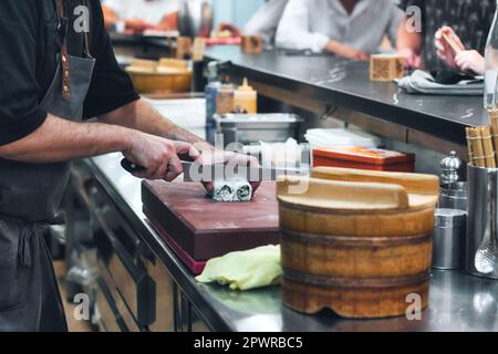 Nahaufnahme der Hände eines Küchenchefs mit einem Messer, der in einer Sushi-Bar eine Sushi-Brötchen mit Reis und Seegras schneidet, mit Menschen im Hintergrund Stockfoto