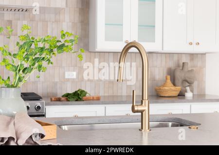 Ein Wasserhahn mit grauer Steininsel, goldenem Wasserhahn, Edelstahlgeräten, braunen Fliesen und weißen Schränken. Stockfoto