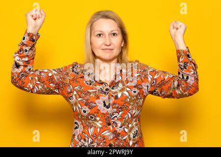 Starke und mächtige weiße Frau, hebt die Arme und zeigt Bizeps, trägt ein gemustertes Hemd, Muster auf gelbem Hintergrund. Sieh dir meine Muskeln an. Stockfoto