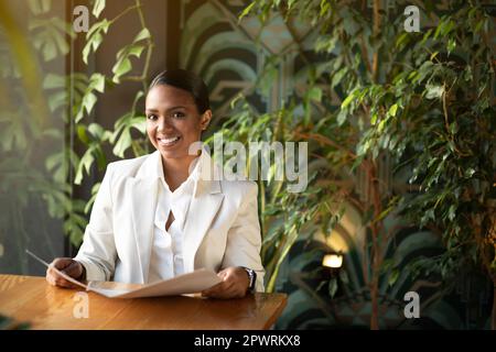 Fröhliche junge schwarze Frau in weißem Anzug, sitzt am Tisch, liest Speisekarte im Öko-Café mit Pflanzen-Interieur Stockfoto