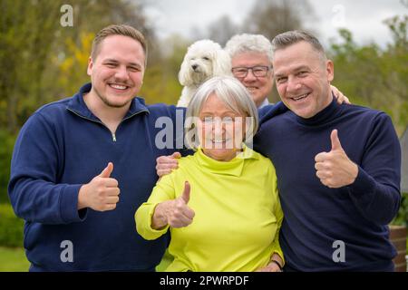 Familie mit Hund, der vor der Kamera fröhlich lacht und als Zeichen des Erfolgs die Daumen nach oben zeigt Stockfoto