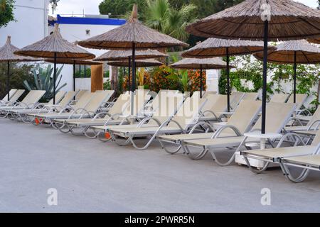 Leere Sonnenliegen, Palmen und Bambusschirme am Pool mit Meer im Hintergrund an einem sonnigen Sommertag Stockfoto