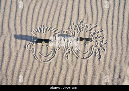 Gesicht im Sand am Strand gezeichnet, mit Sonnenbrille. Sand mit Wellenmuster. Ururlaubsfoto vom Meer Stockfoto