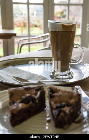 Glasbecher gefüllt mit Latte auf einem Tablett mit Glasfenstern dahinter in einem Café und ein Brownie im Vordergrund Stockfoto