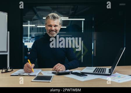 Porträt eines selbstbewussten Geschäftsmanns, Buchhalters, Managers, der im Büro am Tisch sitzt, lächelt und in die Kamera schaut. Arbeiten Sie mit einem Laptop und mit Dokumenten. Stockfoto
