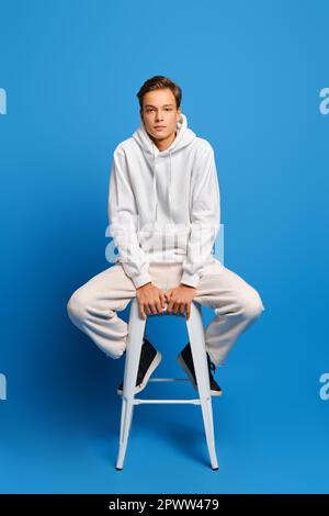 Hübscher Mann mit weißem Sweatshirt und Sweatshirts auf weißem Stuhl über blauem Studiohintergrund sitzend Stockfoto
