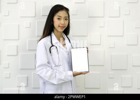 Junger asiatischer Arzt in weißem Kleid und Stethoskop mit weißem Tablet-Computer, der vor einer weißen rechteckigen Keramikwand steht. Stockfoto