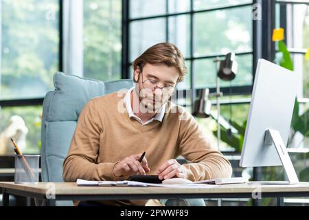 Buchhaltung, Rechnungsprüfung. Ein junger Mann arbeitet mit Dokumenten und Rechnungen im Büro. Berechnet und prüft Berichte, Salden und Budgets auf einem Rechner. Stockfoto