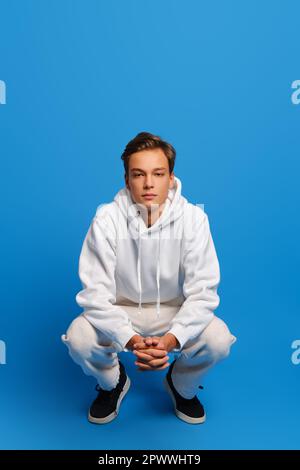 Attraktiver Mann mit weißem Sweatshirt und Sweatshirts hockt auf blauem Studiohintergrund Stockfoto