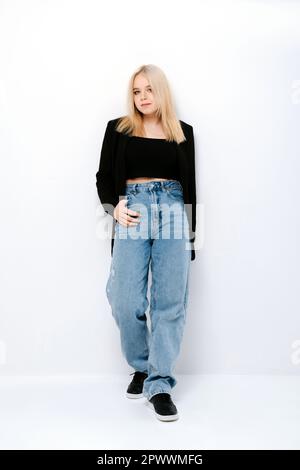 Blondes Mädchen mit schwarzem Oberteil, Jacke und Jeans, die über der weißen Wand posieren Stockfoto