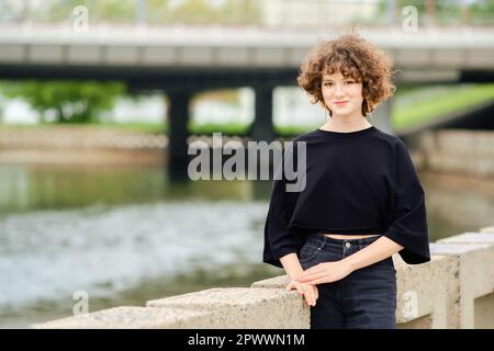 Porträt eines lächelnden Teenagers in schwarzen Jeans und T-Shirt in der Nähe des Flusses Stockfoto