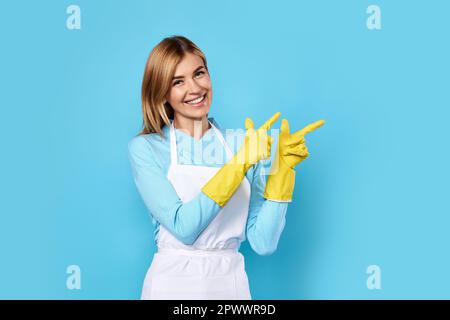 Schöne, lächelnde blonde Frau in gelben Gummihandschuhen, die mit dem Finger auf den Kopierraum zeigt, isoliert auf blauem Hintergrund. Reinigung Stockfoto