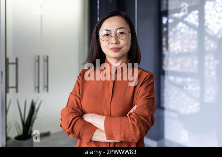 Porträt einer erfahrenen Chefin einer asiatischen Geschäftsfrau, einer Mitarbeiterin, die ernsthaft in die Kamera schaut, einer Frau mit Brille und verschränkten Armen im Büro, die sich auf Erfolg verlassen kann. Stockfoto