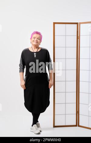Fröhliche reife Frau mit kurzen rosafarbenen Haaren, die in der Nähe eines klappbaren Bildschirms im weißen Raum posiert Stockfoto