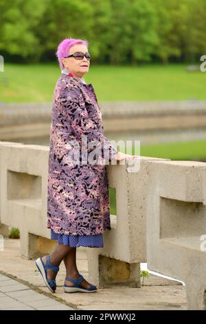 Porträt einer alten Frau mit kurzen rosafarbenen Haaren und Wollmantel, die am Flussufer im Stadtpark steht Stockfoto