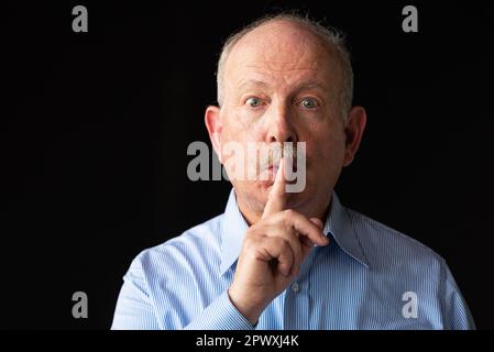 Grauhaariger älterer Mann mit Finger auf den Lippen für Stille oder Geheimnisse vor schwarzem Hintergrund Stockfoto