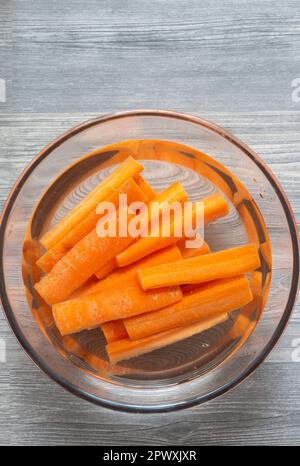 In Scheiben geschnittene Karotten in einer Glasschüssel mit Wasser einweichen. Auf grauem Holzhintergrund Stockfoto
