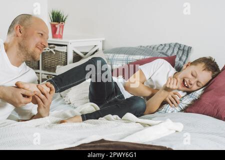 Vater spielt mit seinem Sohn 7-10 auf dem Bett. Papa kitzelt die Füße der Kinder. Familie, die Spaß hat Stockfoto