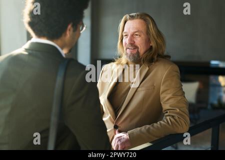 Konzentrieren Sie sich darauf, einen erwachsenen Geschäftsmann zu lächeln, der während der Verhandlung bei einem Arbeitstreffen im Bürozentrum mit einem jungen, ethnischen männlichen Kollegen spricht Stockfoto