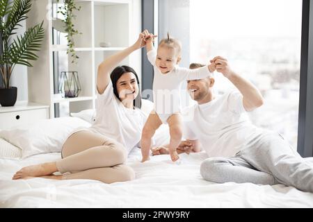 Liebende eltern, die sich an den Händen eines aktiven Mädchens halten und dem Baby helfen, die ersten Schritte auf dem großen weißen Bett zu machen. Fröhliches, lachendes Ehepaar in legerem Outfit, das mit einem fröhlichen Kind spielt. Stockfoto