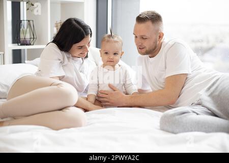 Kaukasische Eltern, die mit dem Baby auf dem Bett liegen und sich von beiden Seiten zärtlich umarmen. Süßes kleines Mädchen mit lustigem Pferdeschwanz, das leicht lächelt und nach vorne schaut. Konzept von Liebe und Familienschätzen. Stockfoto