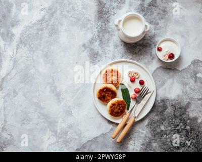Hüttenkäse-Pfannkuchen auf weißem Teller mit Sauerrahm und Beeren auf grauem Hintergrund. Hausgemachte traditionelle ukrainische und russische Syrniki. Gesund Stockfoto