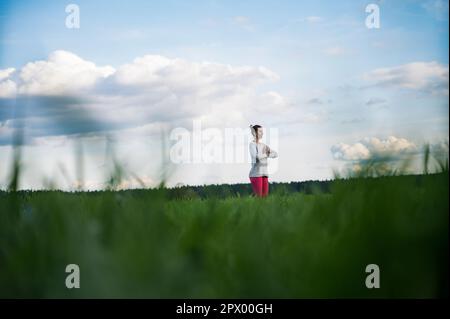 Blick aus tiefem Winkel durch grünes Gras von einer friedlichen jungen Frau, die in der Natur unter bewölktem blauem Himmel steht und mit ihren Handflächen vor ihrem C meditiert Stockfoto