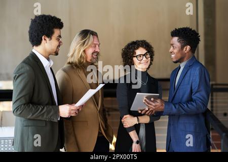 Gruppe glücklicher Broker, die einem jungen afroamerikanischen Kollegen mit Tablet zuhören, der die Schwankungen an den Aktienmärkten präsentiert Stockfoto