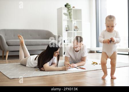 Fröhliche Eltern in lässigen Outfits zeichnen mit bunten Bleistiften, während das niedliche Baby am sonnigen Wochenende im geräumigen Zimmer herumläuft. Junge Erwachsene ermutigen kleine Tochter zu Hause zur Kreativität. Stockfoto