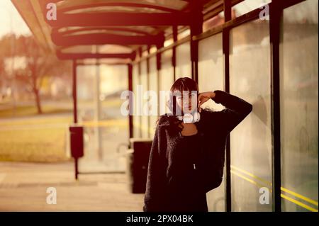 Porträt einer jungen attraktiven Frau in schwarzer Kleidung, die an der Bushaltestelle auf den Bus wartet, urbanes Porträt einer jungen selbstbewussten Frau an der Bushaltestelle, die schwarz trägt Stockfoto