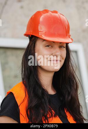 Junge Frau mit rotem Schutzhelm und orangefarbener Warnweste, langes dunkles Haar, schaut in die Kamera und lächelt. Nahaufnahme von Detail zu Gesicht, verschwommene Struktur Stockfoto