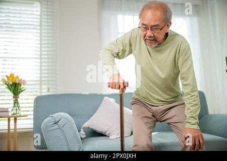 Asiatischer alter Mann mit einer Brille, die mit Gehstock vom Sofa aus aufsteht, ältere Menschen mit Knieschmerzen, die den Griff des Rohrs halten, Senior DIS Stockfoto