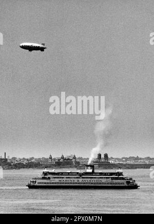 AJAXNETPHOTO. JULI 1975. NEW YORK HARBOUR, USA. - GOODYEAR BLIMP - LUFTSCHIFF SEGELT ÜBER STATEN ISLAND FÄHRE, DIE AUF DEM WEG NACH BATTERY, MANHATTAN IST, WÄHREND SIE DAS ALTE ELLIS ISLAND EINWANDERUNGSZENTRUM (HINTERGRUND) PASSIEREN. FOTO: JONATHAN EASTLAND/AJAX REF:232404 113 Stockfoto