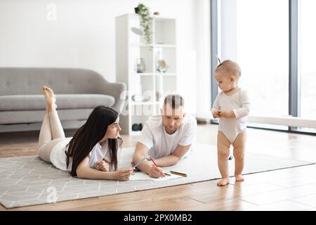 Fröhliche Eltern in lässigen Outfits zeichnen mit bunten Bleistiften, während das niedliche Baby am sonnigen Wochenende im geräumigen Zimmer herumläuft. Junge Erwachsene ermutigen kleine Tochter zu Hause zur Kreativität. Stockfoto