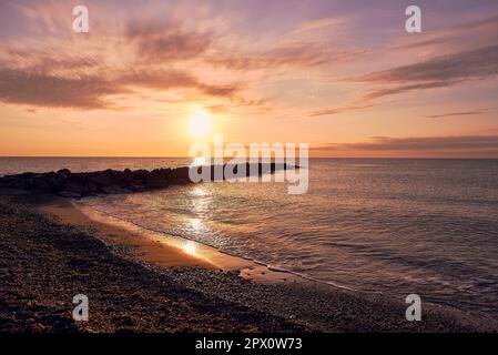 Sonnenaufgang am Kiesstrand mit Wellenbrecher. Unendlicher Hintergrund, orange und rötliche Farben, leerer Strand, kleine Steine. Moncofar Stockfoto