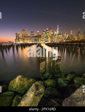 Blick auf Lower Manhattan in der Dämmerung von Brooklyn aus. Der Rest eines alten Piers ist im Vordergrund und die Skyline der Stadt im Hintergrund zu sehen Stockfoto