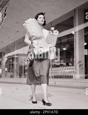 1950S FRAU, DIE ZWEI GROSSE PAPIERTASCHEN MIT LEBENSMITTELN VOR DEM SUPERMARKT MIT SICH FÜHRT - S6332 HAR001 HARS ZUFRIEDENHEIT WEIBLICHE HEIM LEBEN KOPIERRAUM VOLLE LÄNGE DAMEN PERSONEN SHOPS B&W SHOPPER BRÜNETT HOMEMAKER SHOPPERS HOMEMAKERS LOW ANGLE HOUSEWIVES LÄDEN ELEGANT LEBENSMITTELGESCHÄFT VOR PAPIERTASCHEN, HANDEL, JUNGE ERWACHSENE FRAU, SCHWARZ-WEISSE GESCHÄFTE, KAUKASISCHE ETHNISCHE HERKUNFT, HAR001 HIGH HEELS ALTMODISCH Stockfoto