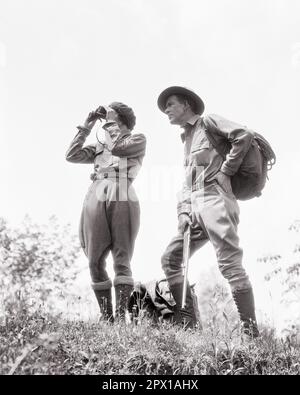 1920S JAGDPAAR MANN MIT RUCKSACK, FRAU MIT GEWEHR, DIE EIN FERNGLAS BENUTZT UND KLASSISCHE OUTDOOR-SPORTKLEIDUNG TRÄGT STIEFEL - H3403 HAR001 HARS FITNESS STYLE GESUNDE TEAMARBEIT WAFFEN LIFESTYLE FRAUEN VERHEIRATET LÄNDLICHER EHEPARTNER EHEMÄNNER GESUNDHEIT NATUR KOPIEREN WELTRAUM FREUNDSCHAFT VOLLE LÄNGE DAMEN PERSONEN GEWEHR MÄNNCHEN HUNTER CONFIDENCE HIKE B&W JAGD PARTNER FREIHEIT ZIELE VISION AKTIVITÄT PHYSISCHES ABENTEUER ENTDECKUNGSKRAFT HIKER LOW WINKELSPORT MIT FLEXIBILITÄTSMUSKELN SCHUSSWAFFEN WANDERER JAGEN JODHPURS MITTELERWACHSENER MANN MITTELERWACHSENER FRAU ZUSAMMENSEIN EHEFRAUEN RUCKSACK SCHWARZ UND WEISS Stockfoto