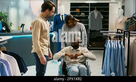 Männlicher Rollstuhlfahrer, der trendige Kleidung in einem Modeboutique-Laden einkauft und Waren an Kleiderbügeln ansieht. Person mit chronischer Behinderung, die moderne formelle Kleidung kauft, kleines Unternehmen. Stockfoto